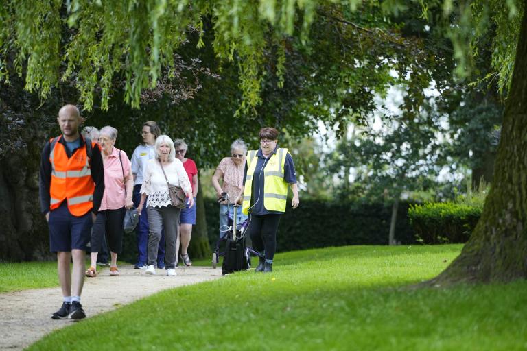 A male adult walking outdoors wearing a high-vis jacket followed by a group of mixed adults