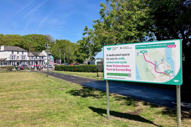 A sign welcoming people to a new cycling and walking route. A cycle path running through a park in the background.