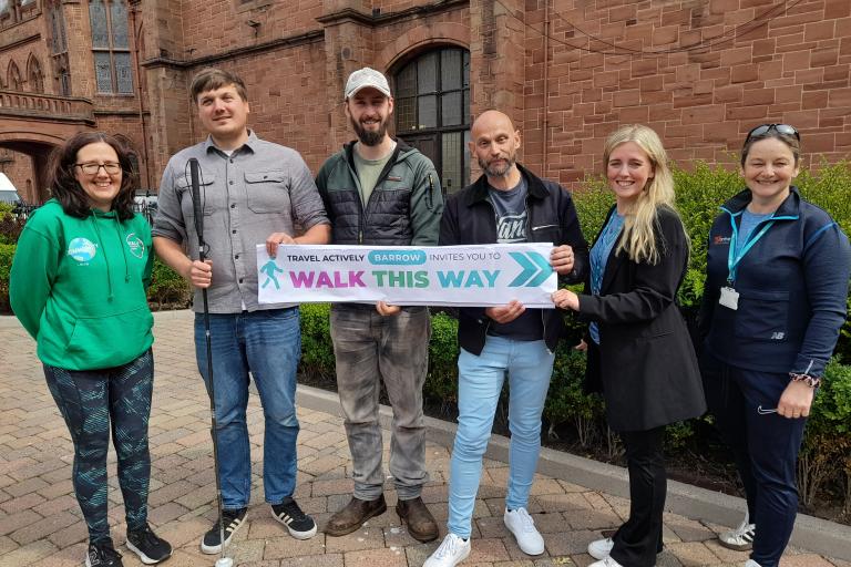 a group of mixed adults in a group smiling at the camera. They are holding a banner which reads: Travel Actively Barrow invites you to Walk This Way