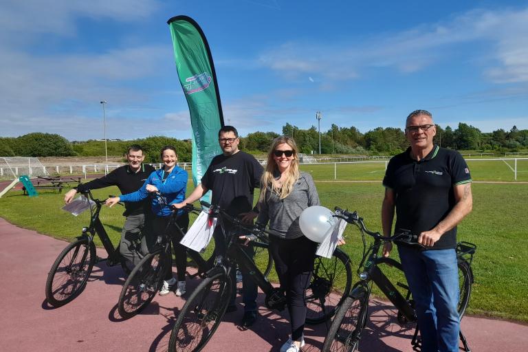 A group of adults stood outdoors next to e-bikes. They are smiling at the camera