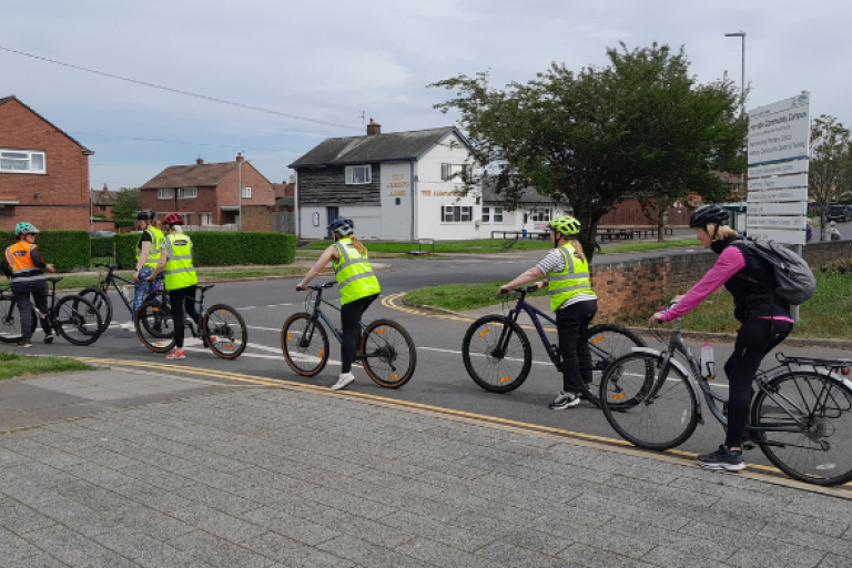 A group of cyclists on a road. They are stopped at a junction and are wearing high-vis jackets