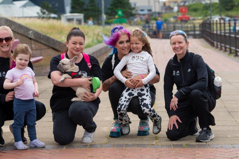 A group of ladies, children and a dog kneeling outdoors whilst on a walk and smiling at the camera