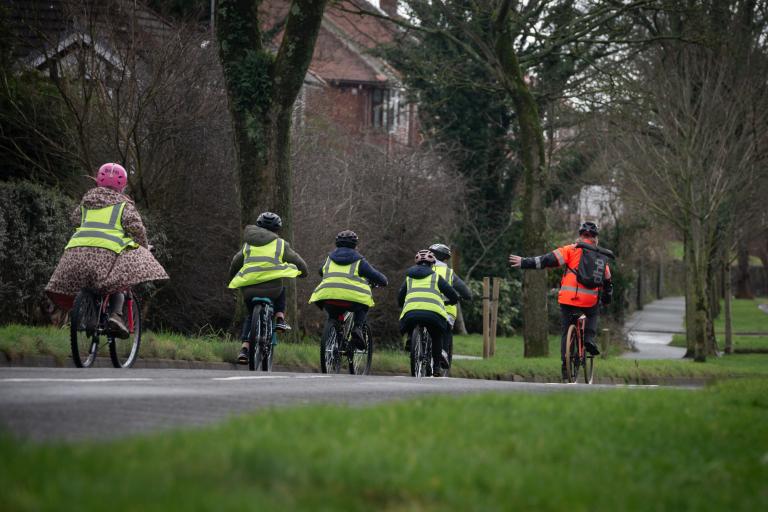 Five children wearing green high-vis jackets cycling along a road. Infront is a cycling instructor signalling left he is wearing an orang high-vis jacket. In the background are trees and grass verges. 