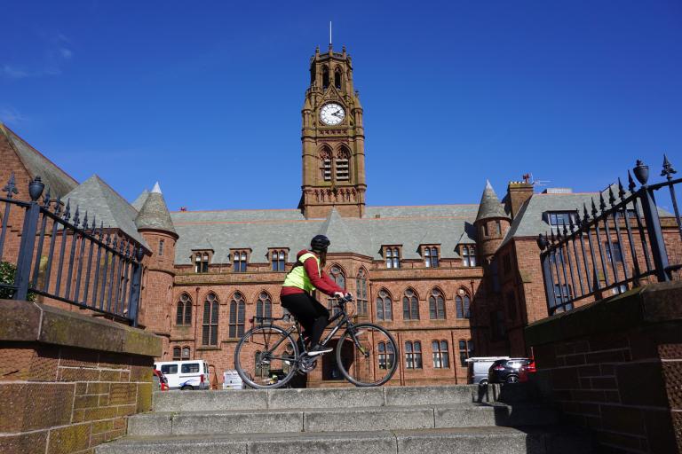 a lday riding a bike wearing a yellow high vis jacket.  Behind her is blue sky and Barrow Town Hall and Clock