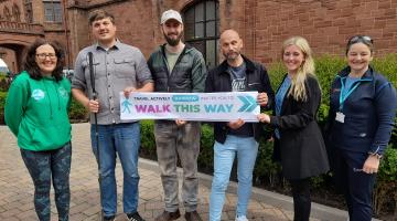 a group of mixed adults in a group smiling at the camera.  They are holding a banner which reads: Travel Actively Barrow invites you to Walk This Way 
