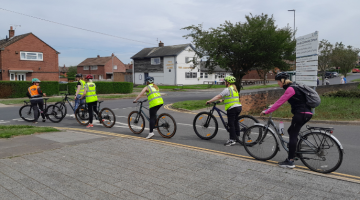 A group of cyclists on a road.  They are stopped at a junction and are wearing high-vis jackets
