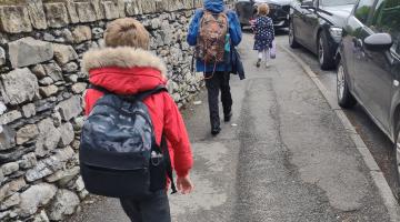Three children walking to school.  Image shows the children from behind. They are carrying school backpacks.