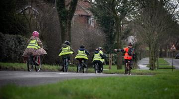 Five children wearing green high-vis jackets cycling along a road. Infront is a cycling instructor signalling left he is wearing an orang high-vis jacket. In the background are trees and grass verges. 