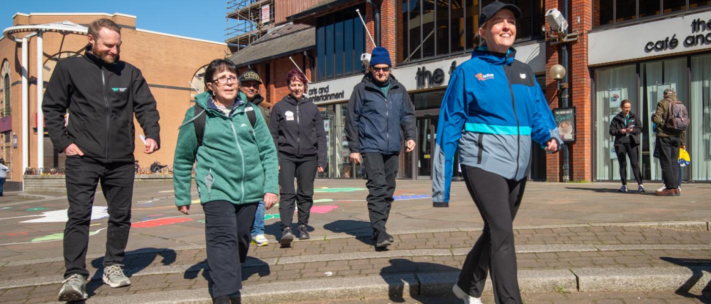 A group of mixed adults walking through Barrow town centre in the sunshine.